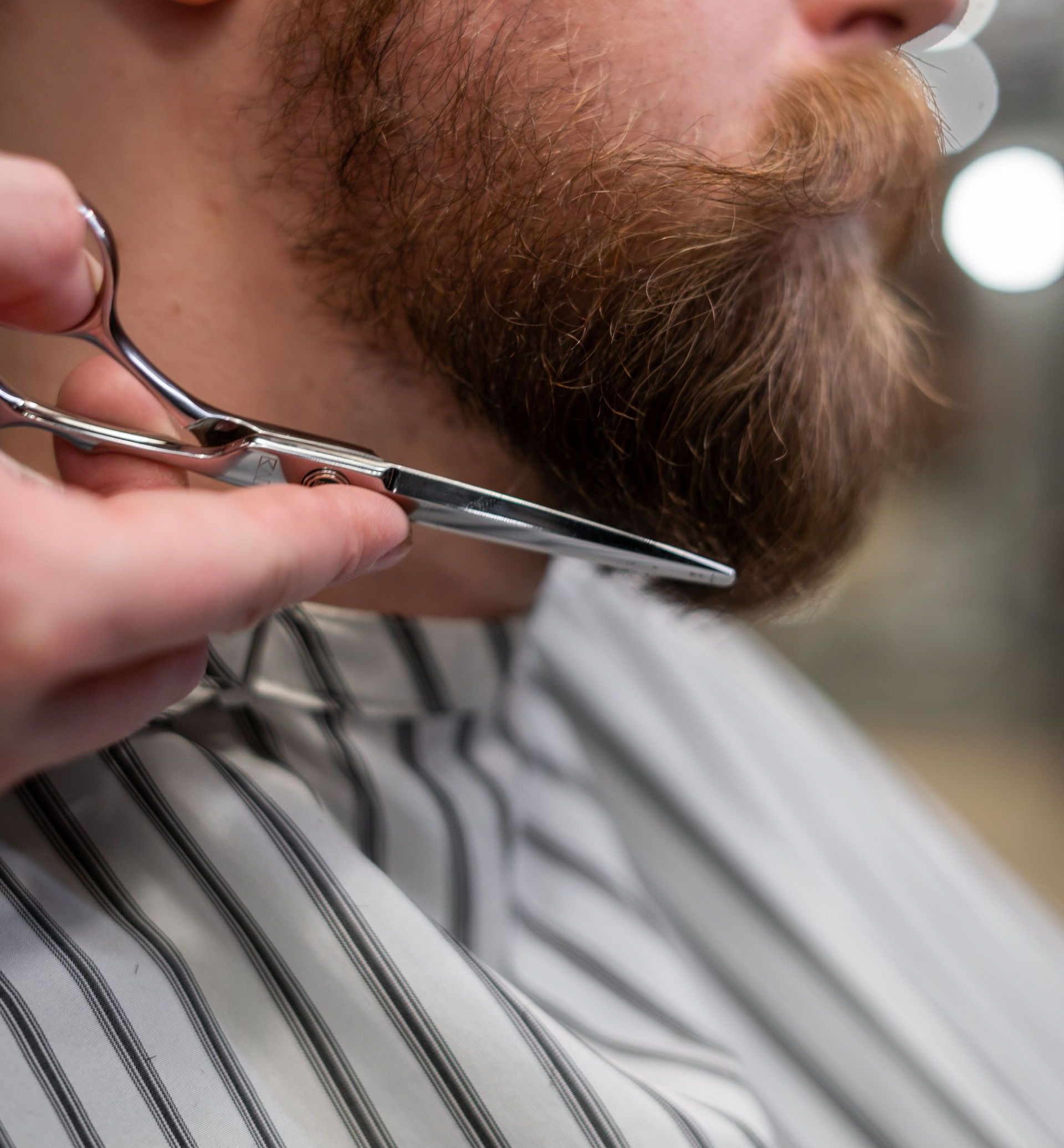 A professional barber trims a bearded man's beard with scissors in a barbershop.